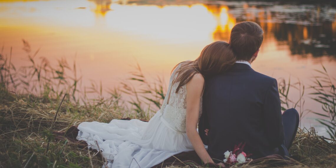 wedding couple sitting on green grass in front of body of water at sunset